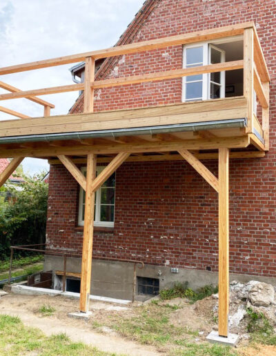 Holzbalkon auf Stützen an einem Backsteinhaus.